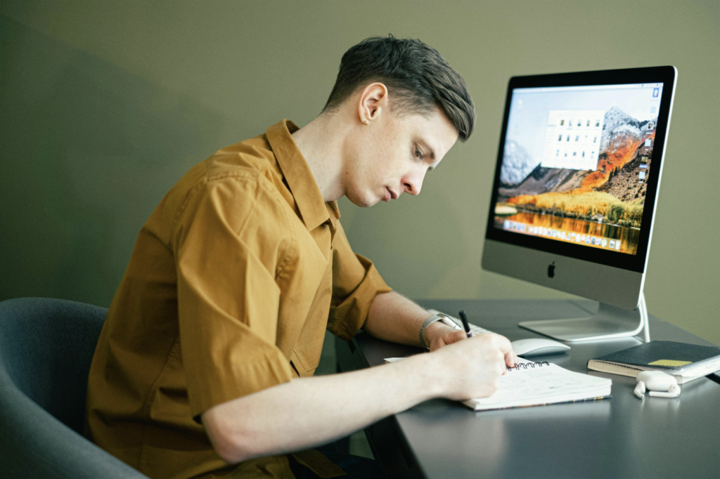 Young man sitting at his desk by a computer taking notes